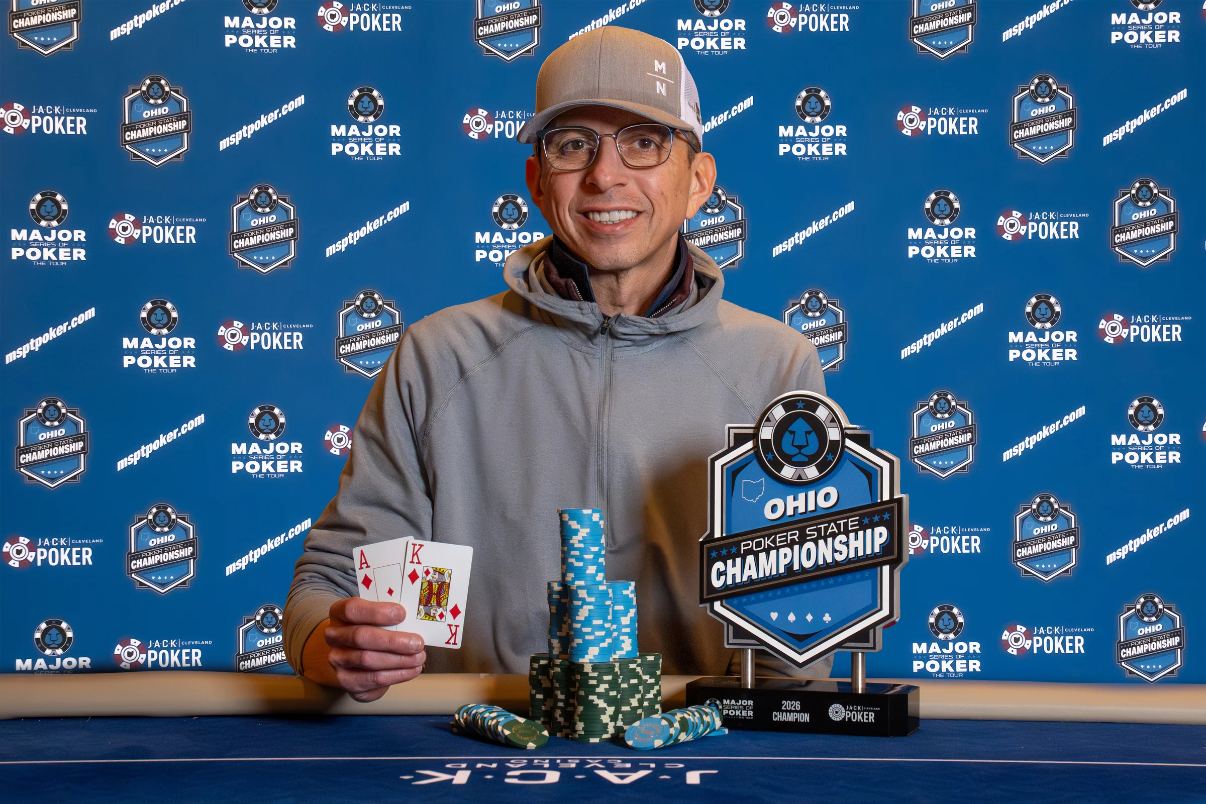 A tournament champion holds ace-king beside chips and an Ohio Poker State Championship trophy.