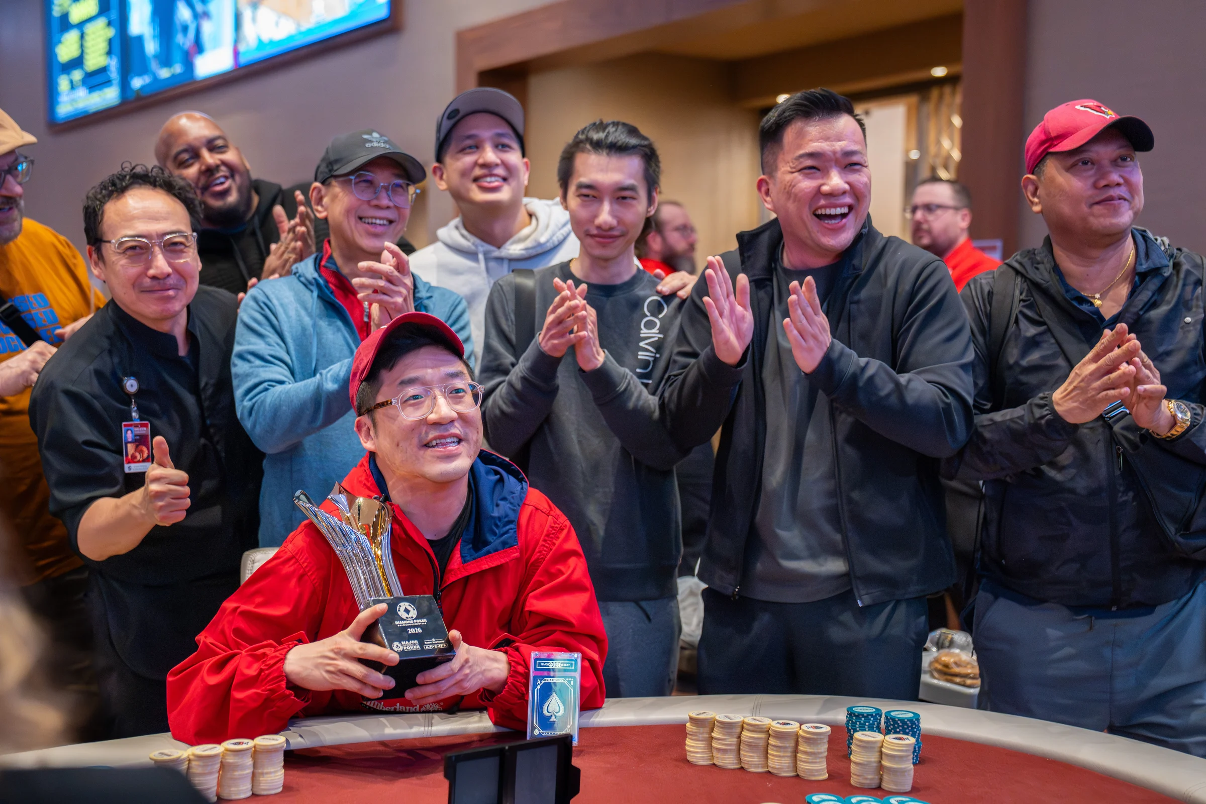 A seated card tournament player holds a trophy while a group applauds behind him.