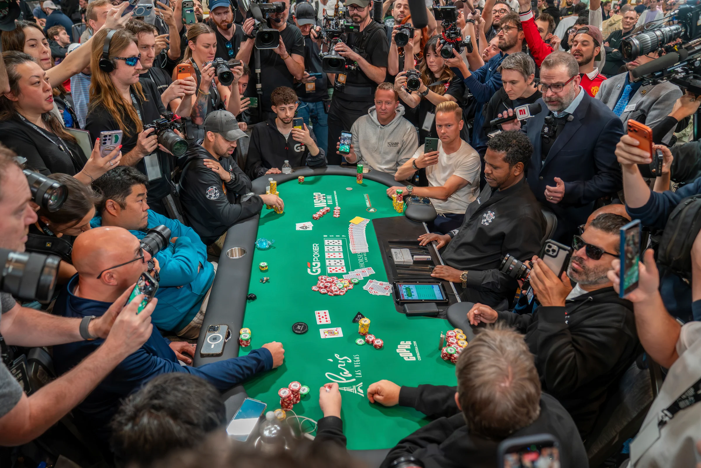 poker players sit at a crowded televised table surrounded by photographers and spectators.