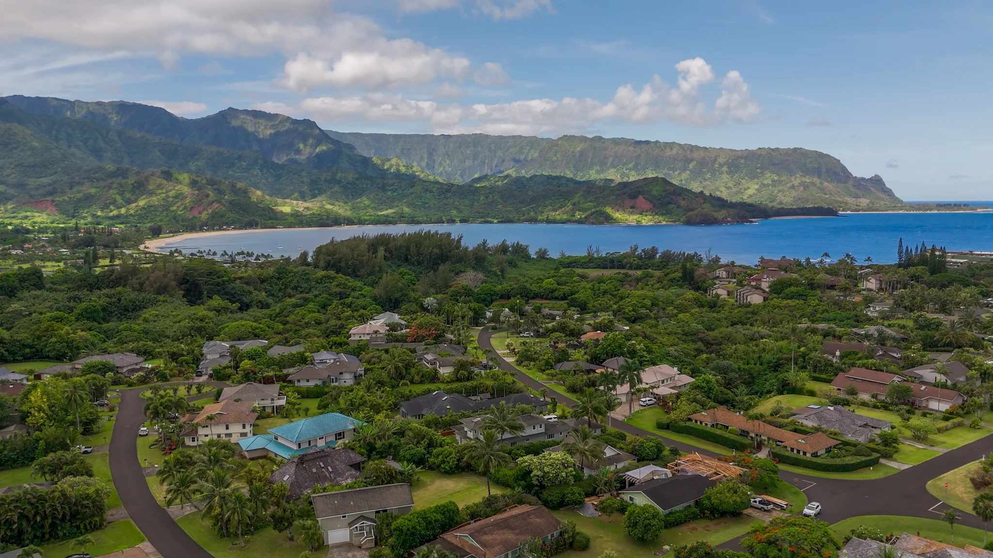Aerial panorama of a coastal neighborhood, blue lagoon, and green mountain ridge beneath cloudy skies.