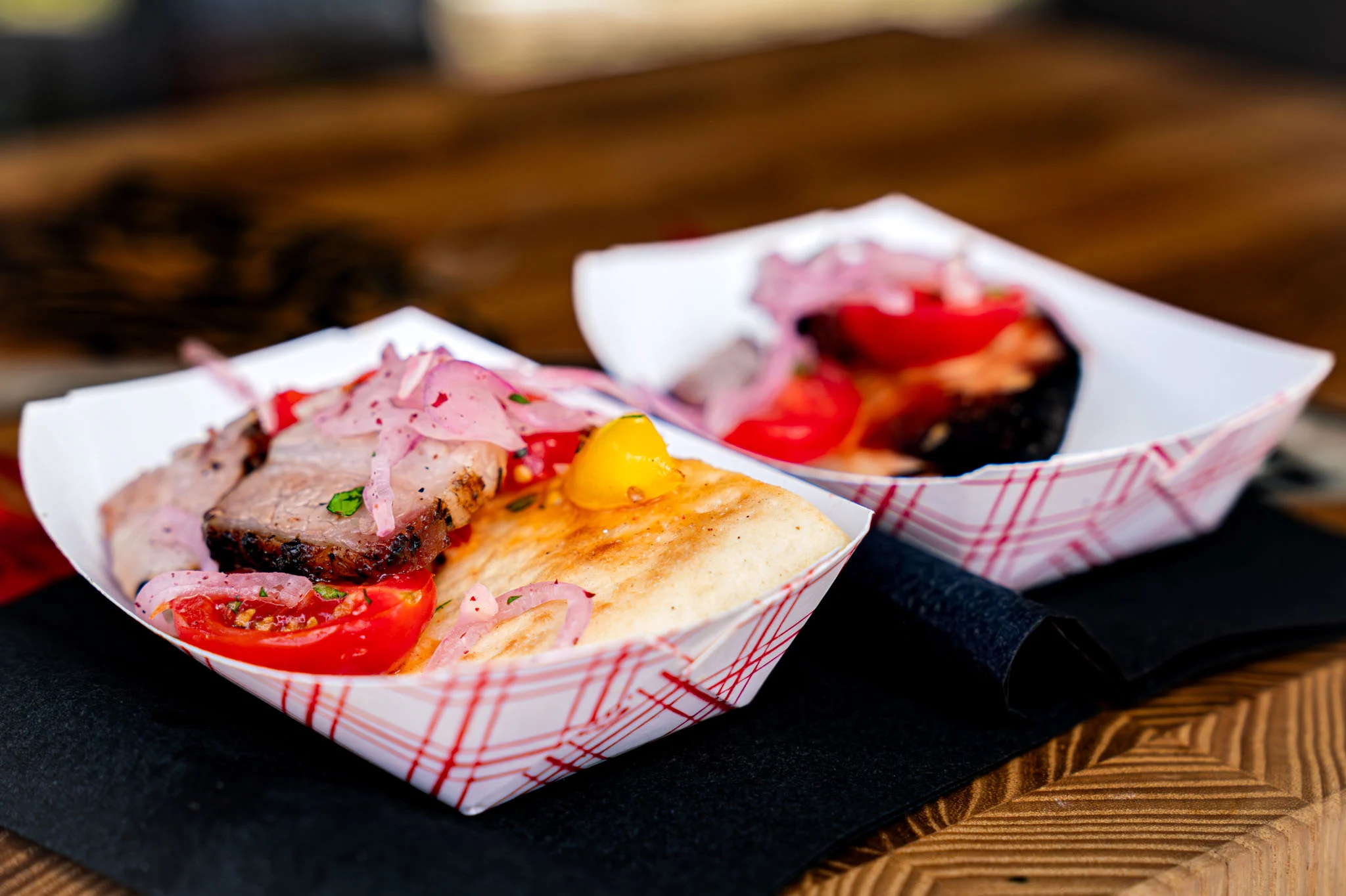 Two barbecue tasting trays hold sliced meat, tomatoes, pickled onions, and grilled flatbread.