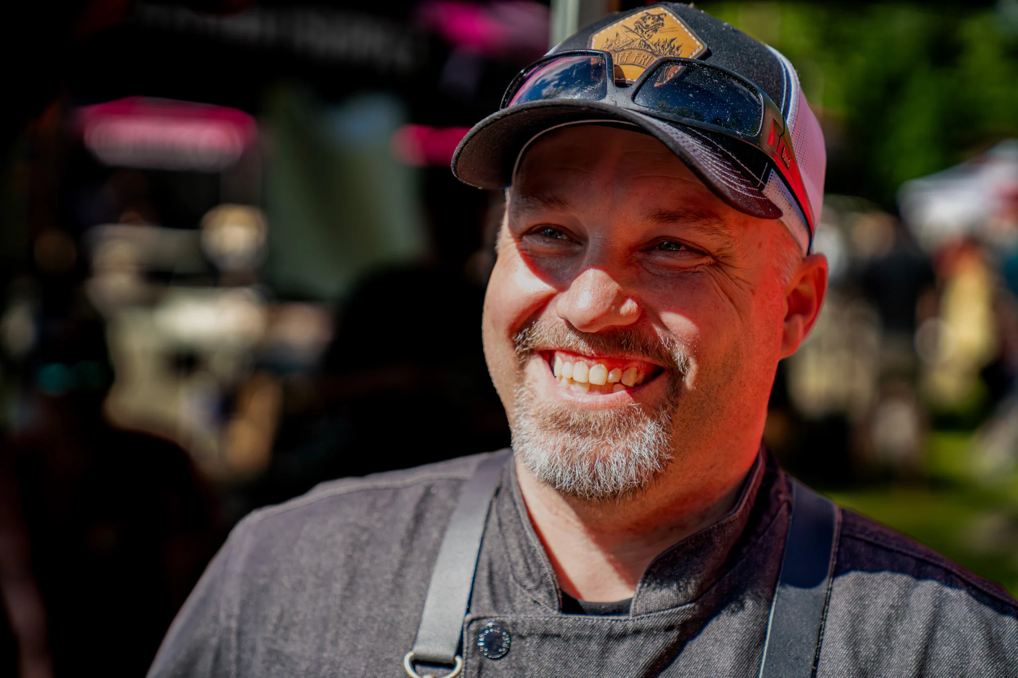 A smiling cook in a cap and apron poses at an outdoor barbecue event.