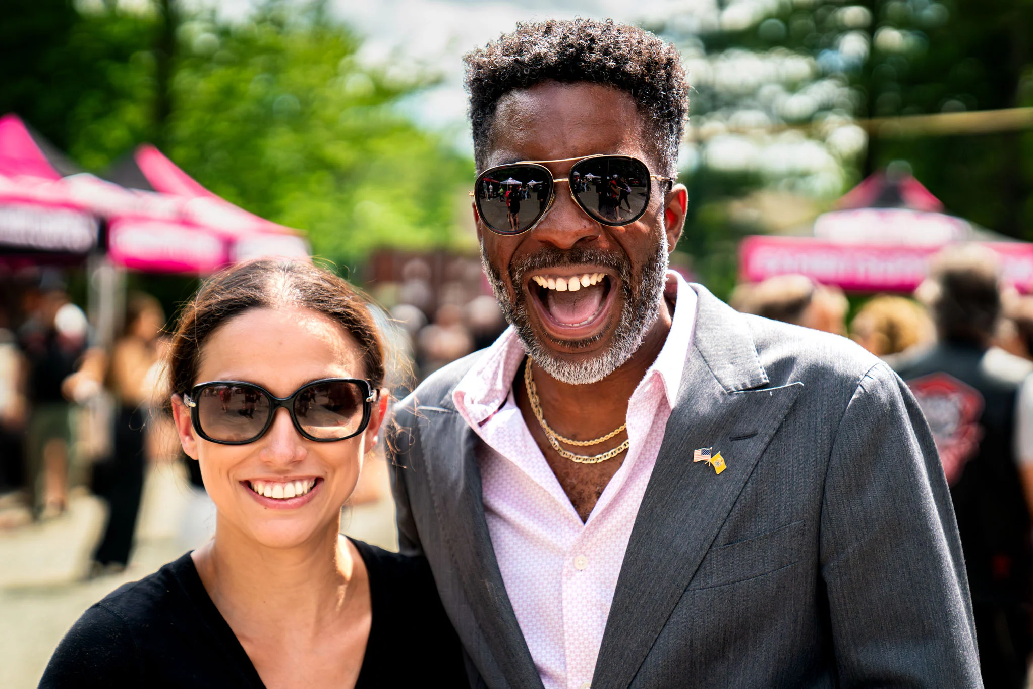 Two guests in sunglasses smile together amid tents and crowds at an outdoor festival.