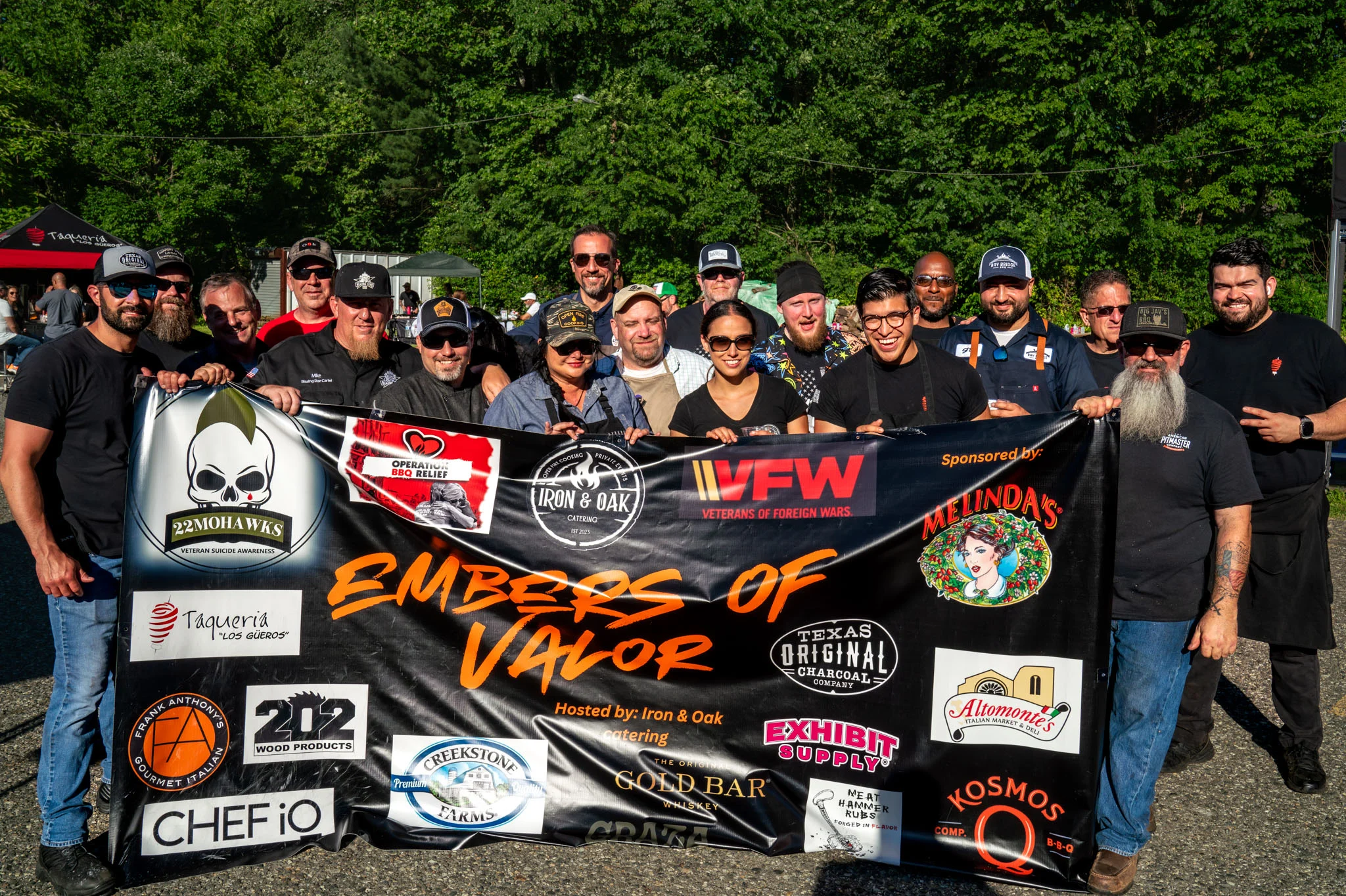 A large group poses behind an Embers of Valor banner at an outdoor barbecue event.
