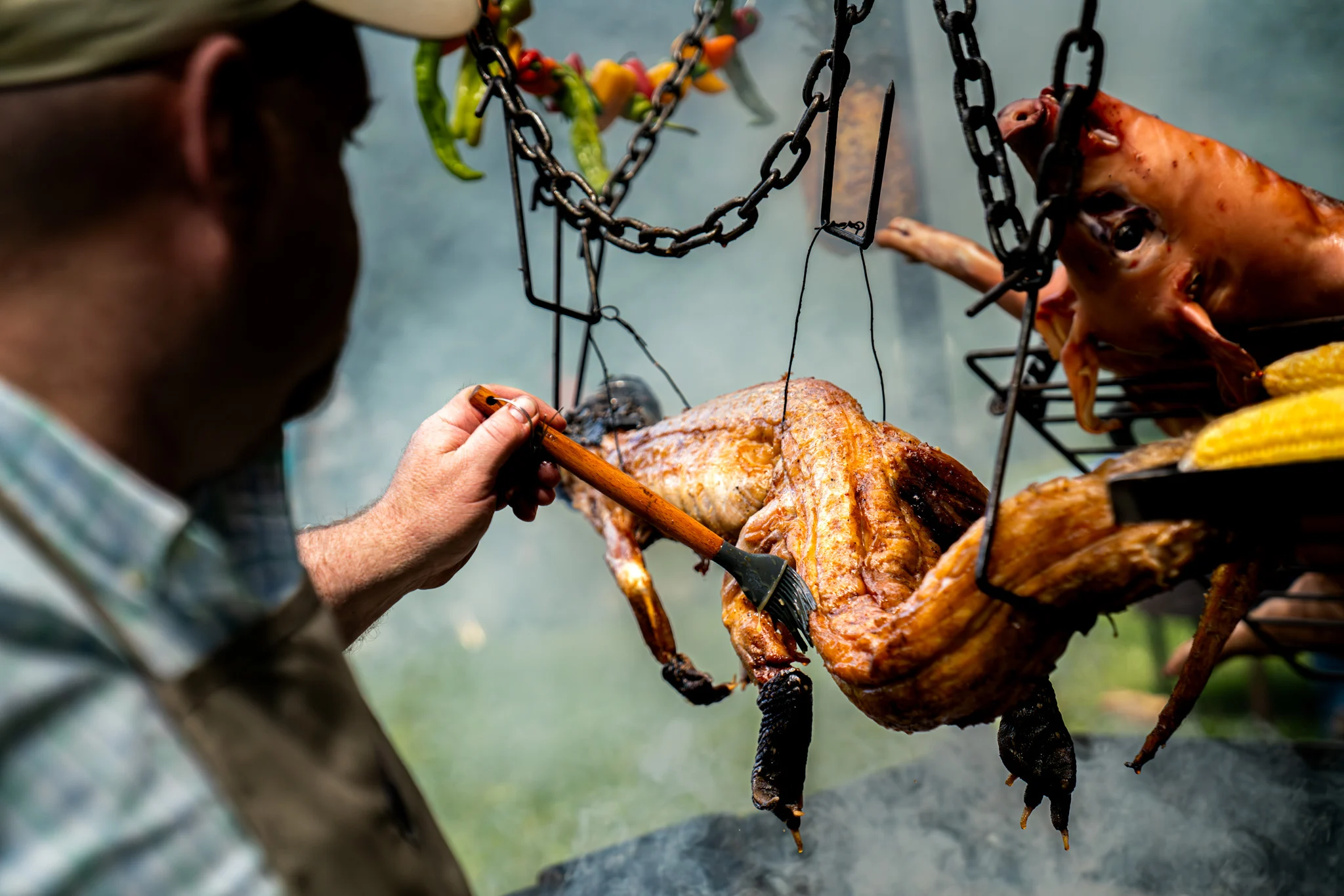 A cook brushes glaze onto hanging roast poultry beside a pig roast and corn.