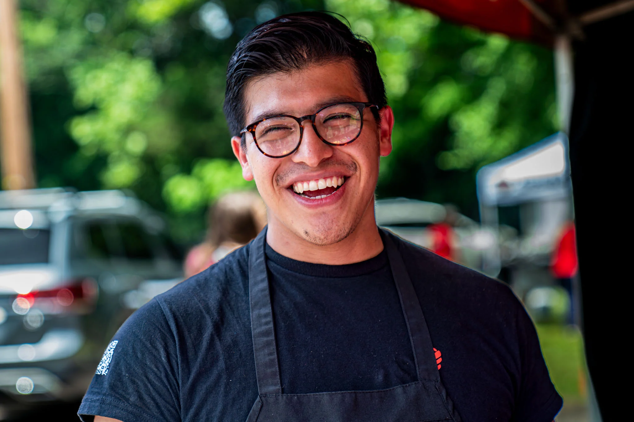 A smiling cook wearing glasses and a black apron stands at an outdoor barbecue event.