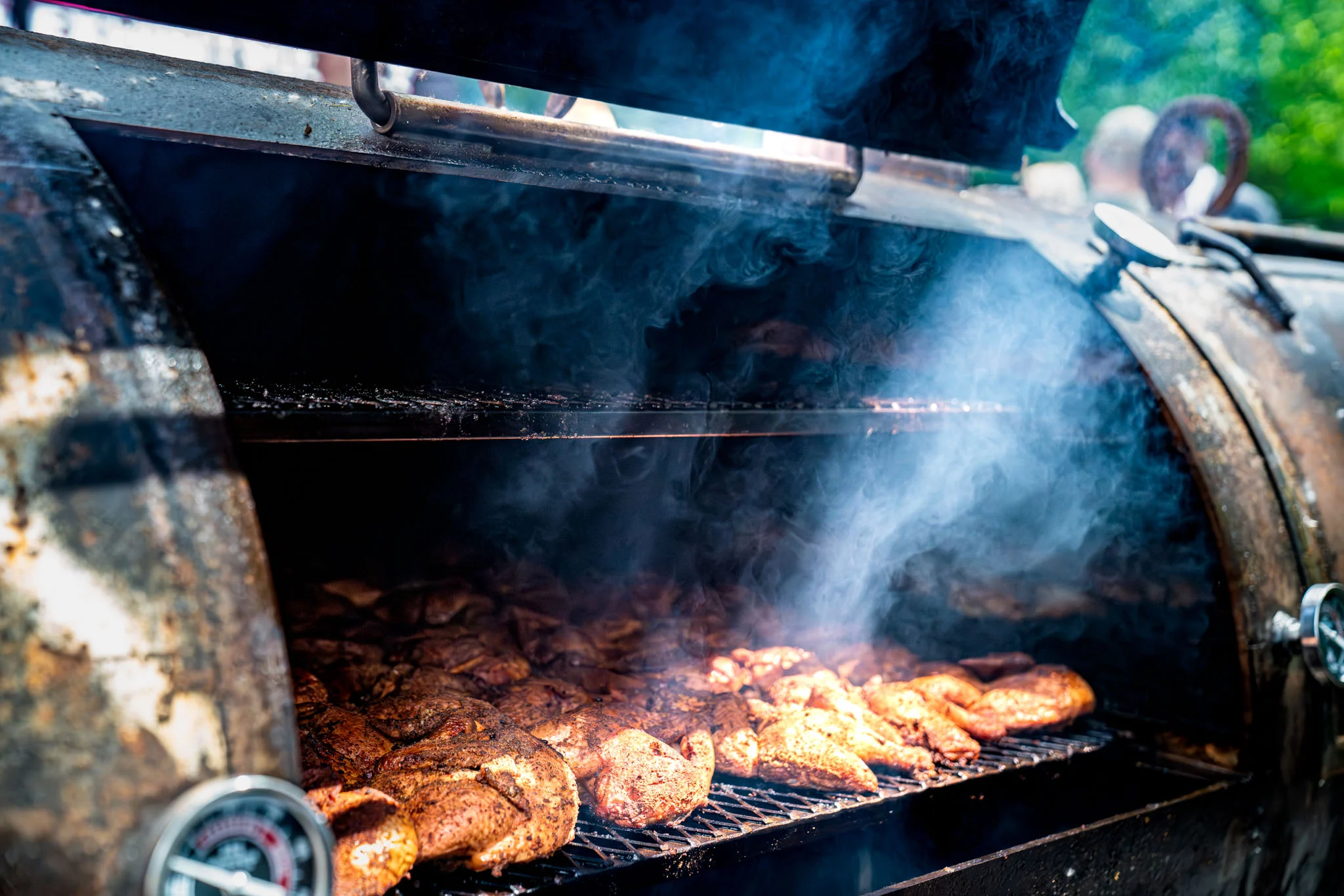 Seasoned poultry cooks inside a smoking barrel grill at an outdoor barbecue event.