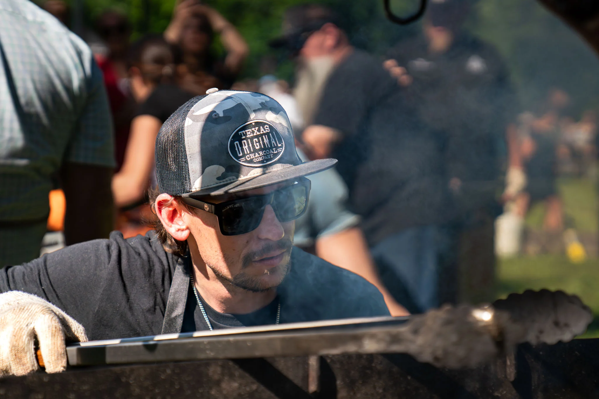 A cook wearing sunglasses tends a smoky grill during an outdoor barbecue event.