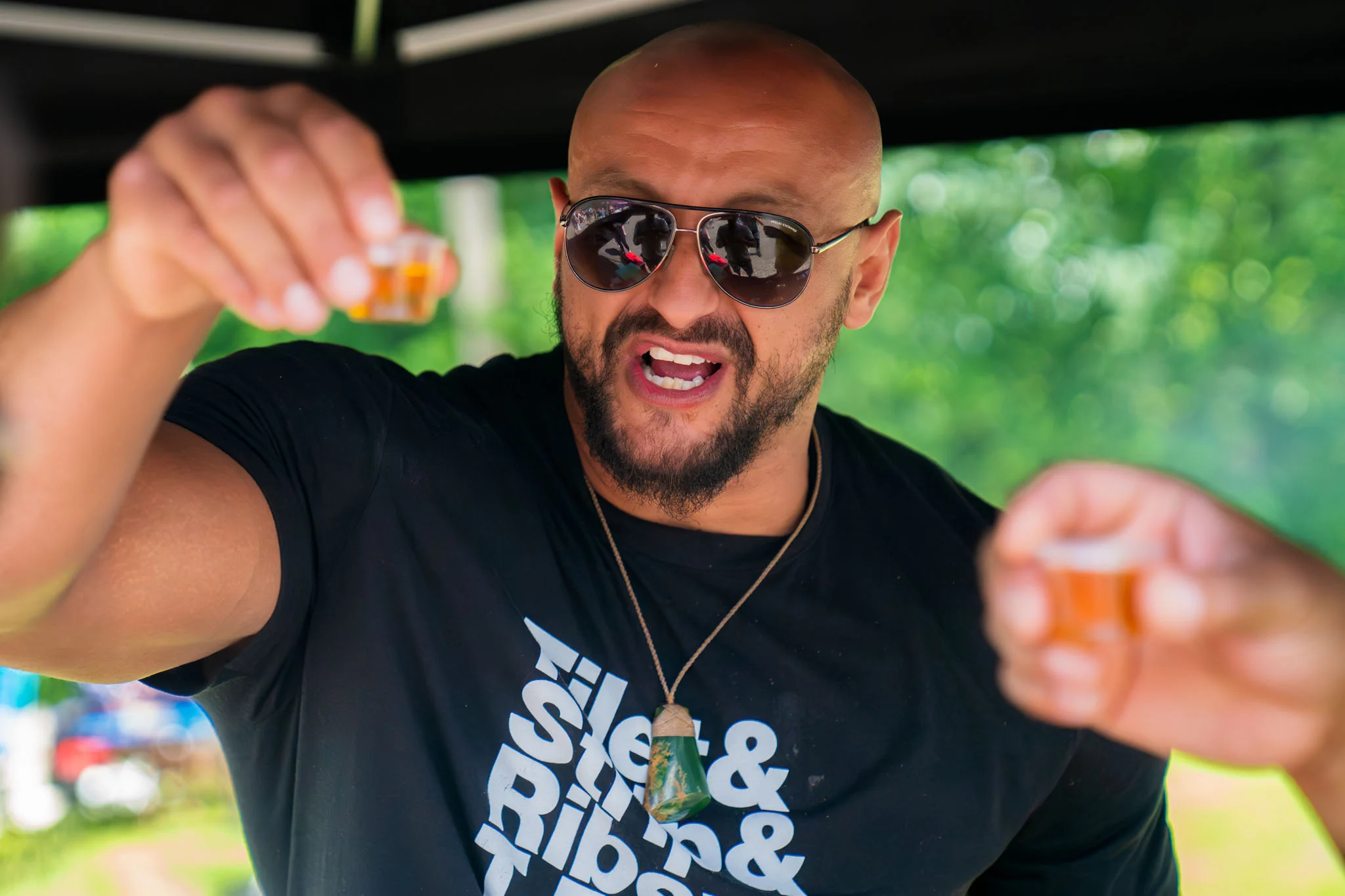 A man in sunglasses raises two small tasting cups beneath an outdoor event tent.