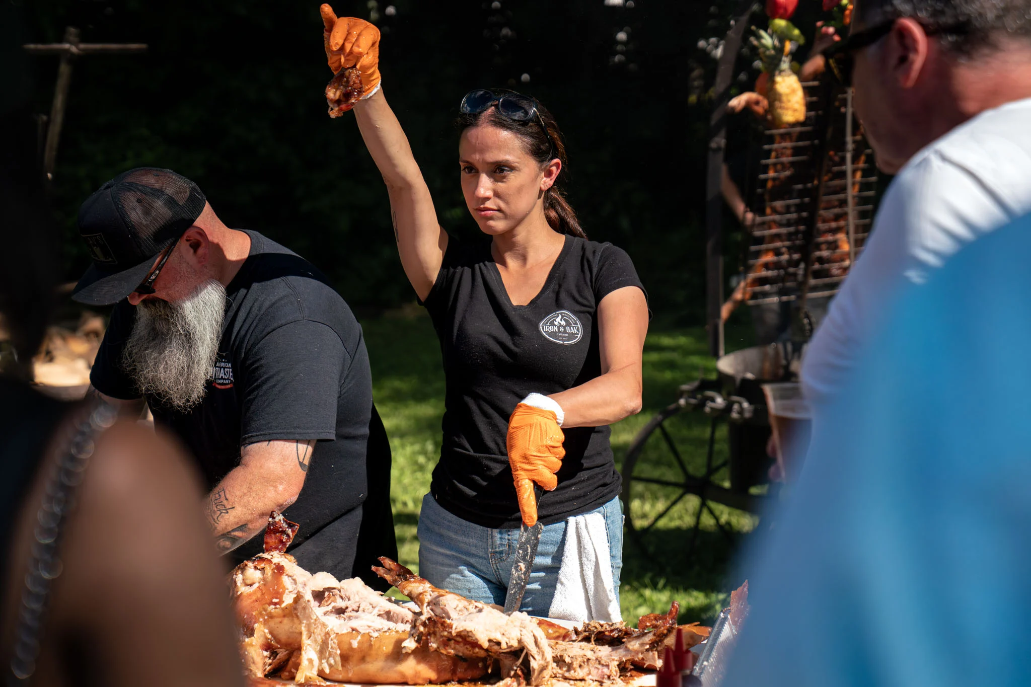 Two cooks carve a whole roasted pig while preparing food for an outdoor crowd.