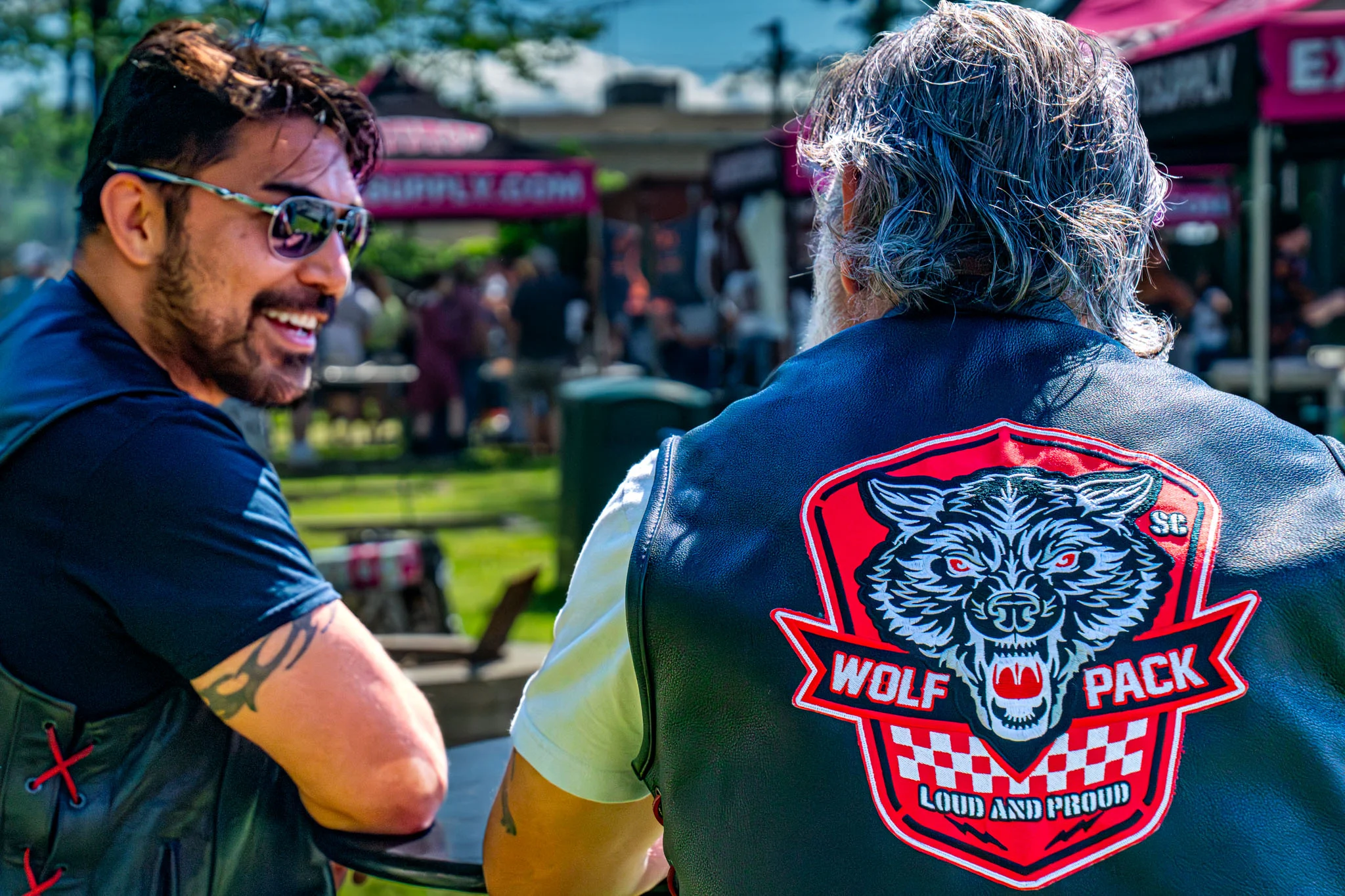 Two men talk beside cooking stations, one wearing a Wolf Pack leather vest.
