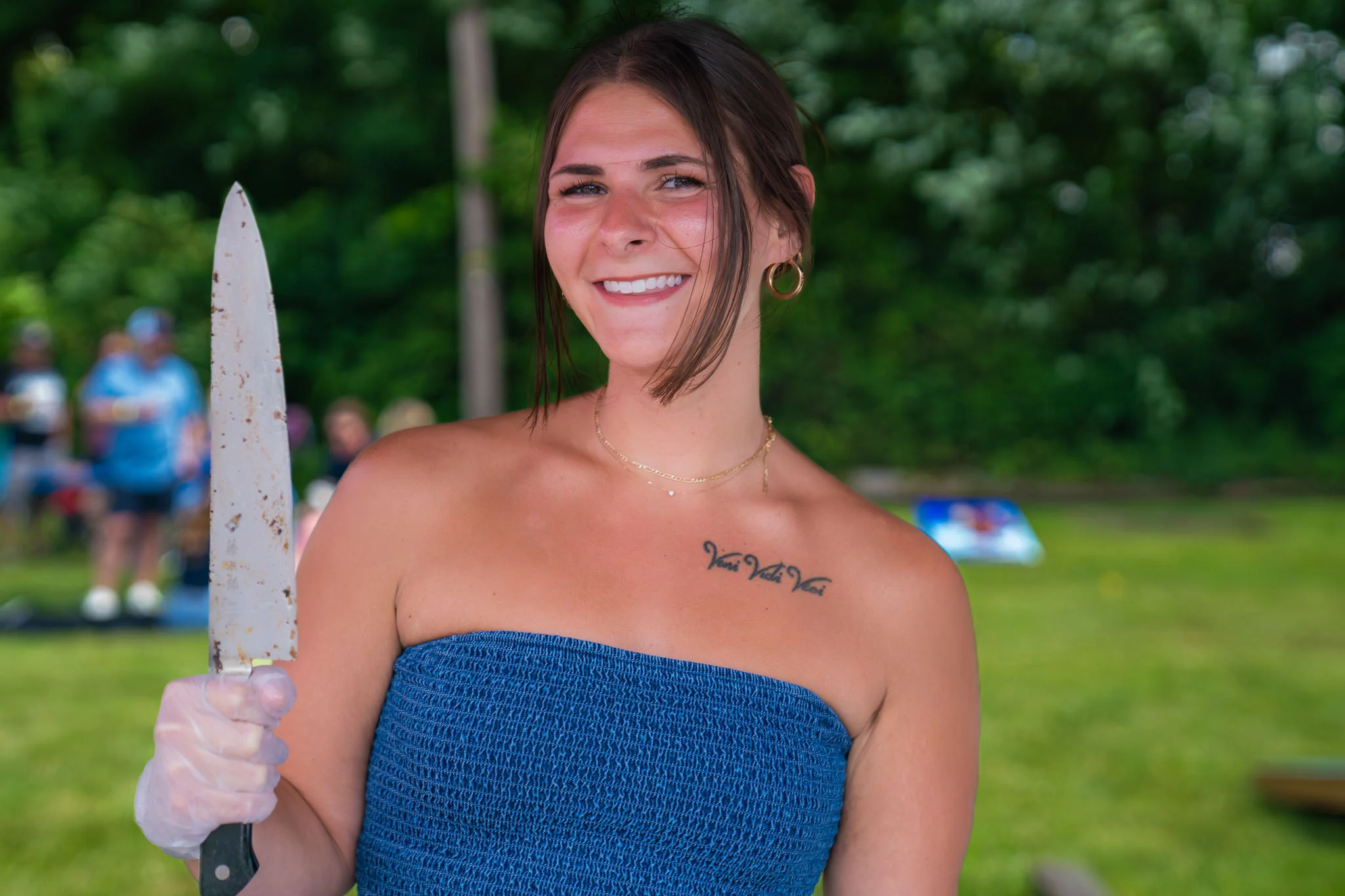 A smiling woman wearing a food-service glove holds a large chef's knife outdoors.