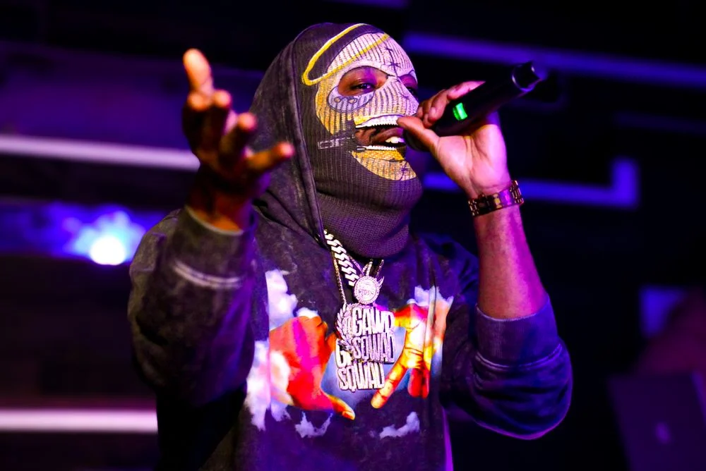 Masked performer reaching toward the audience under purple light