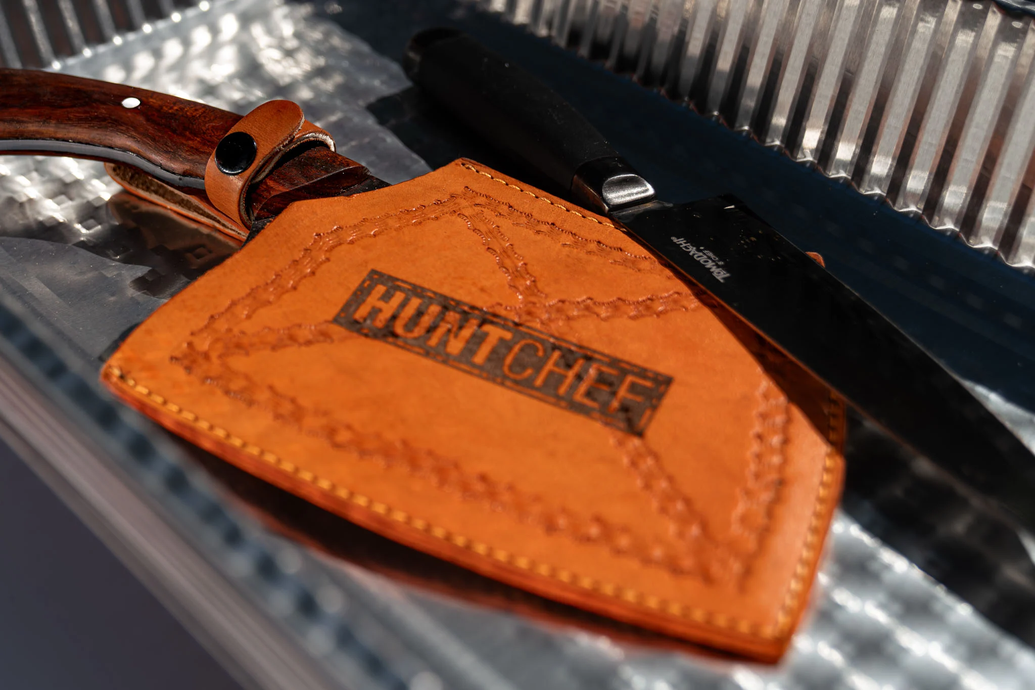 A HuntChef leather knife sheath rests beside cooking knives on a metal prep surface.