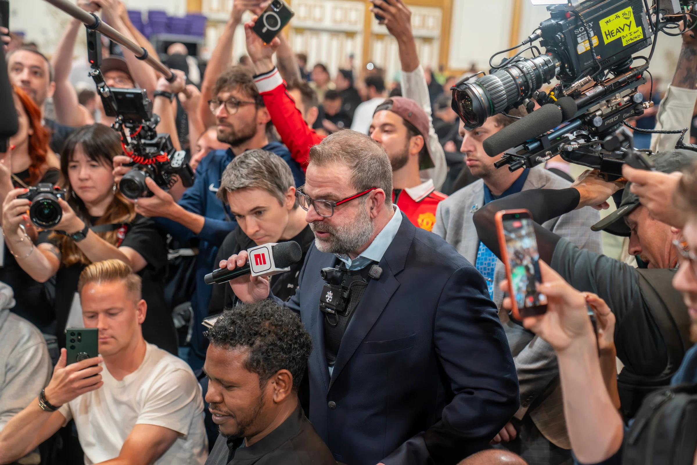 Media cameras crowd around a suited speaker holding a microphone during a poker event.