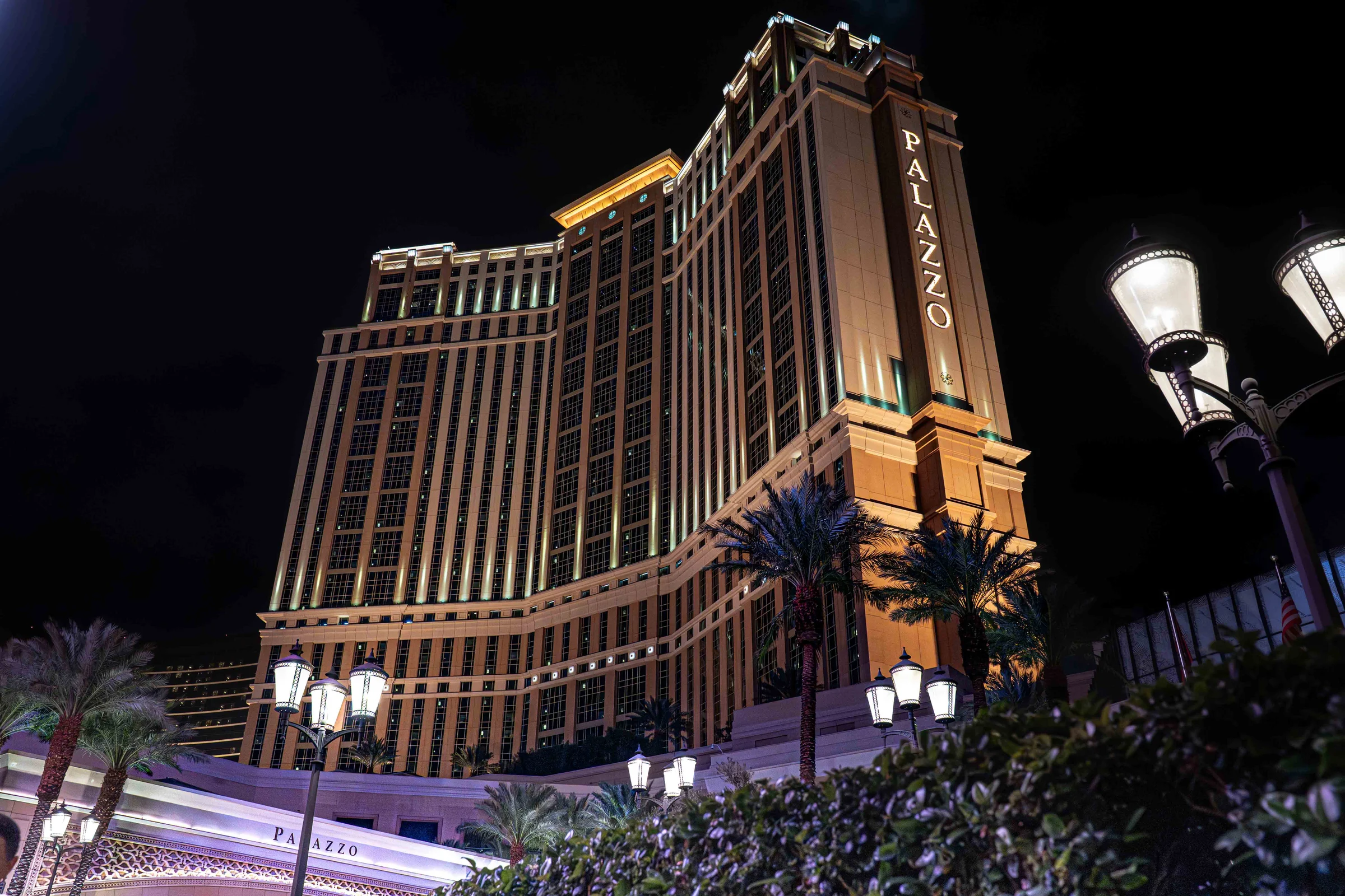 The Palazzo hotel tower rises above palm trees and glowing streetlamps at night.