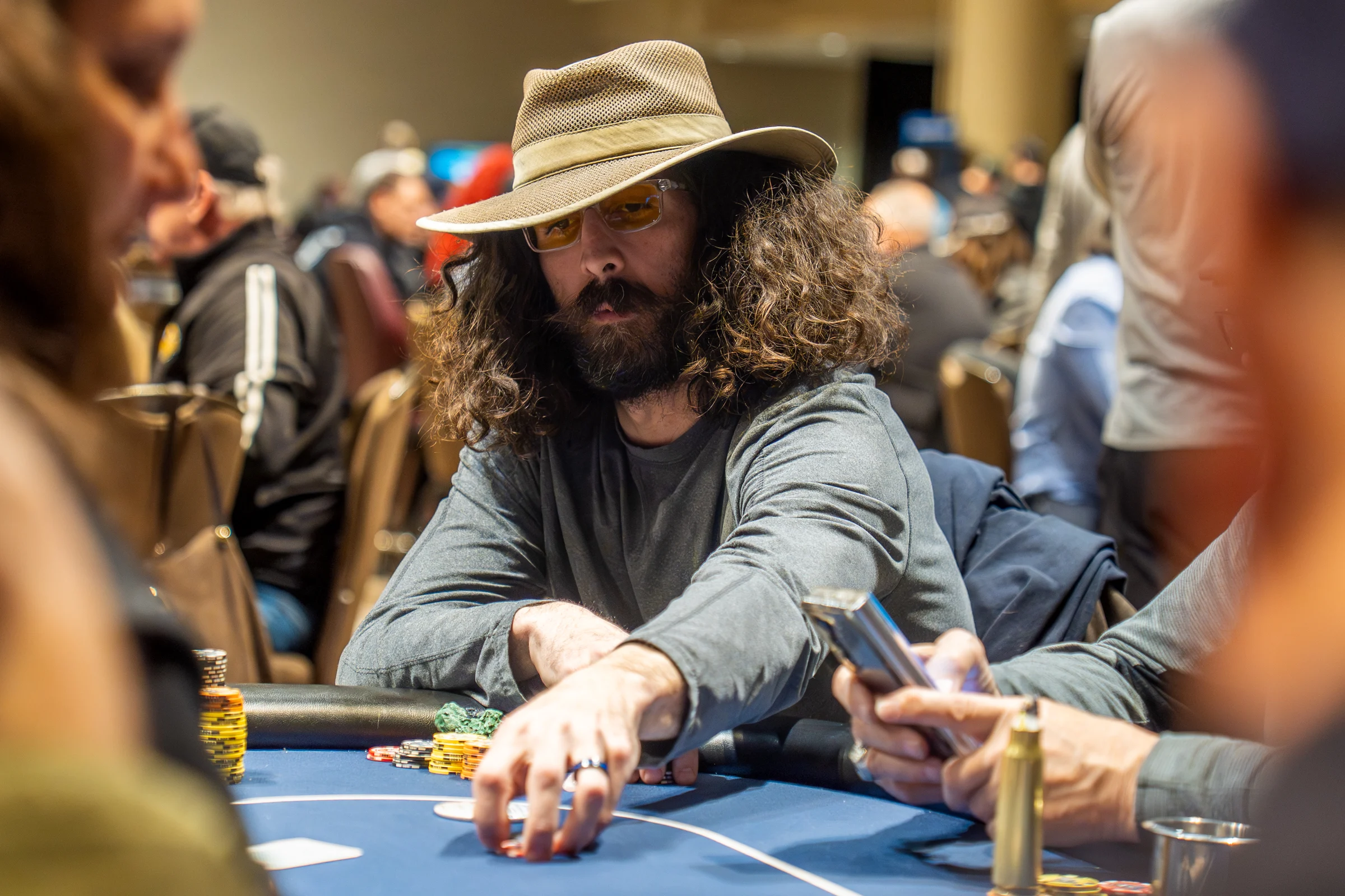 A bearded poker player in a wide-brim hat reaches toward chips at a tournament table.