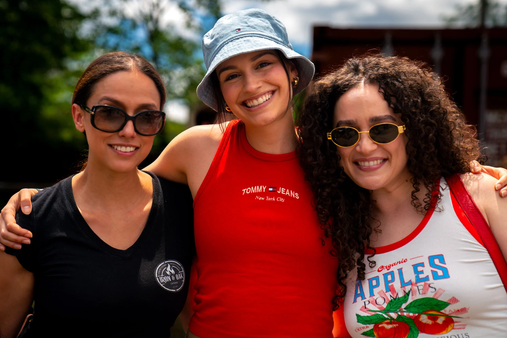 Three women wearing summer clothes and sunglasses pose together outdoors in bright sunlight.
