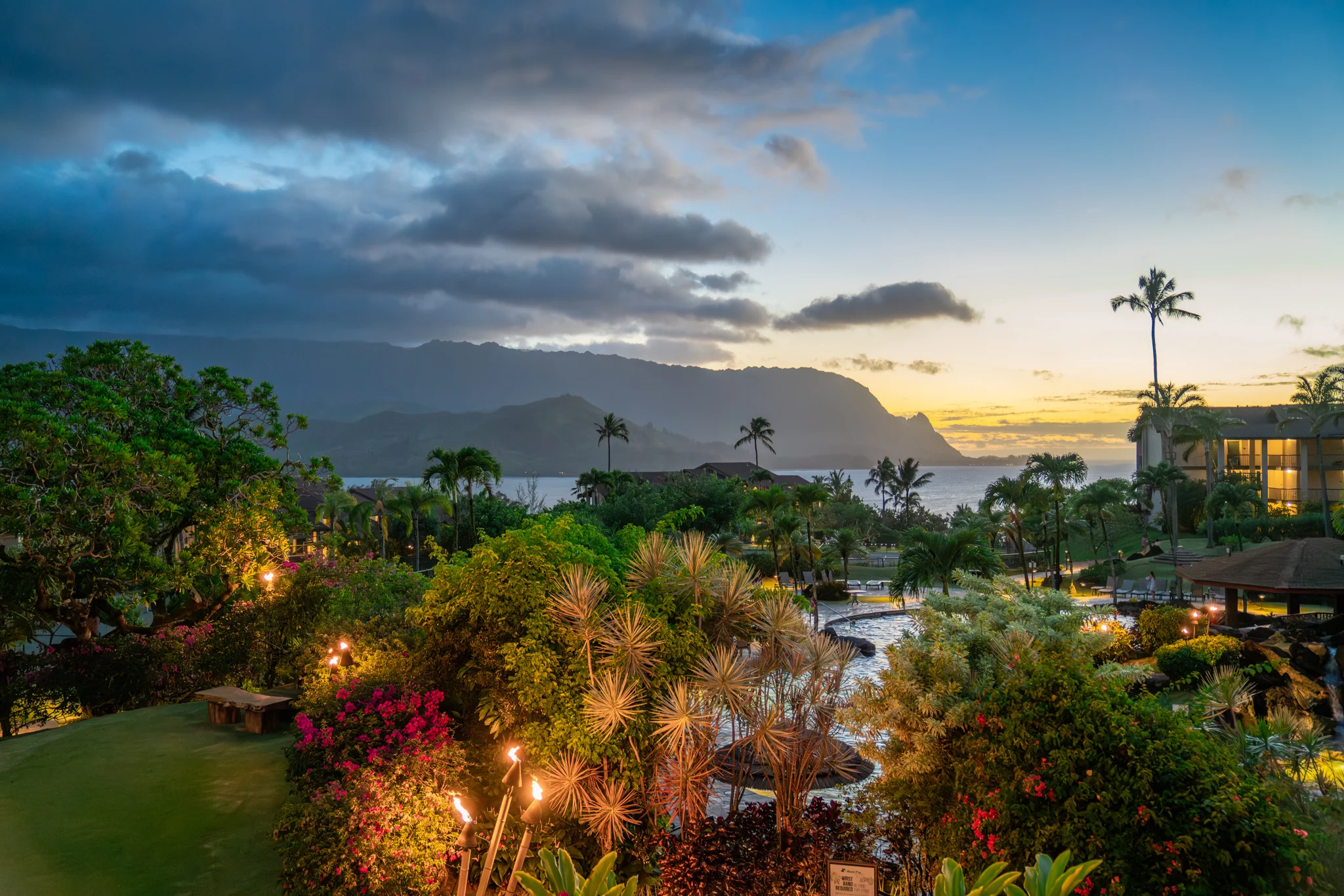 A tropical resort garden overlooks a bay and mountain ridge beneath a glowing sunset.